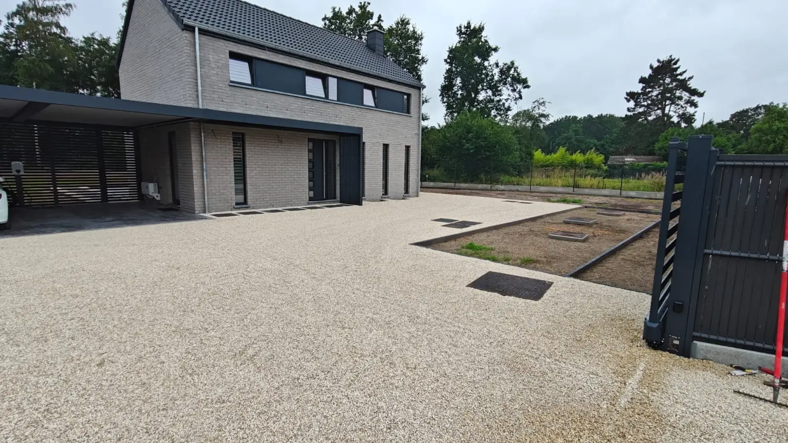 Allée en gravier beige devant une maison en briques grises avec carport, portail coulissant anthracite à droite et massifs de terre au fond