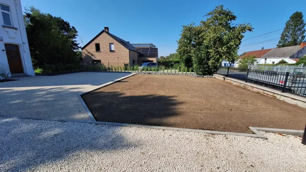 Allée en gravier clair terminée devant une maison blanche, avec à côté une grande surface de terre nivelée bordée d'une bordure