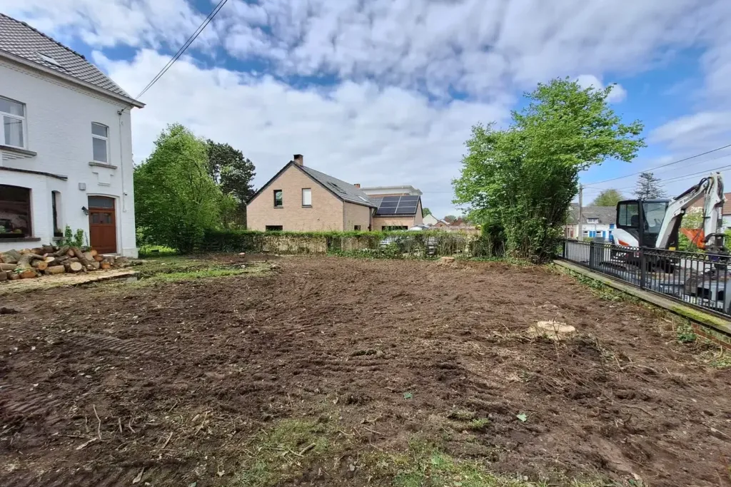 Terrain de terre nivelée devant une maison blanche, avec une mini-pelle à l'arrêt près de la clôture au fond
