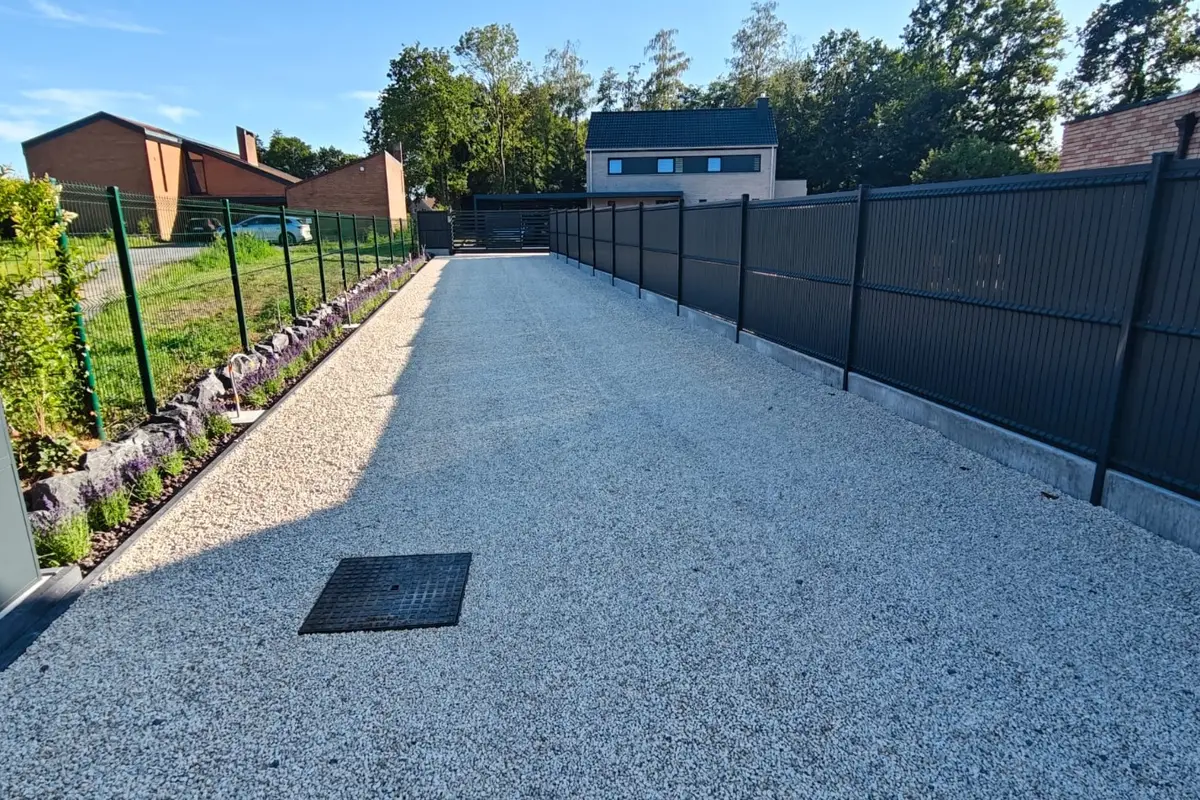 Longue clôture à panneaux anthracite le long d'une allée en gravier clair, avec une plate-bande du côté gauche et un regard en fonte au sol