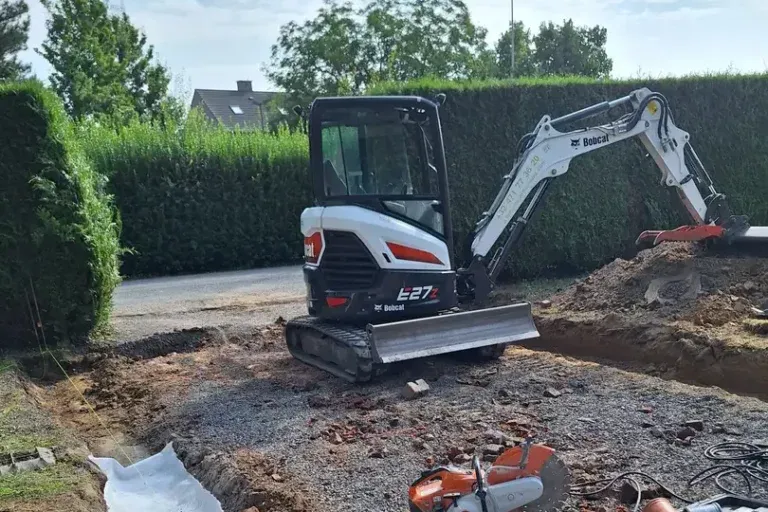 Mini-pelle à l'arrêt à côté d'une tranchée garnie de géotextile blanc, avec une meuleuse et une mallette d'outils sur le gravier