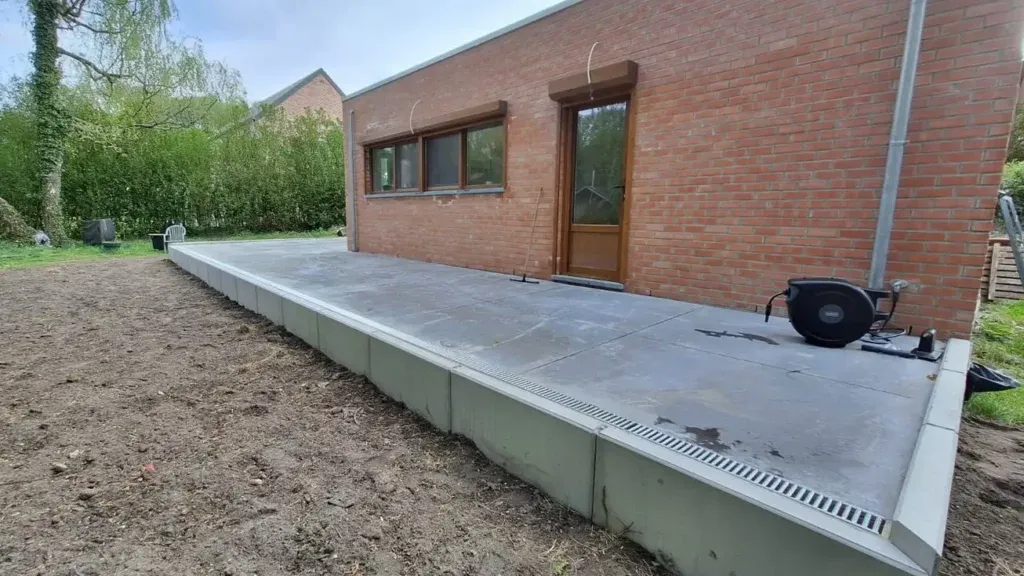 Grands éléments de béton préfabriqué gris en forme de L alignés le long d'une dalle, avec un caniveau à grille sur le dessus, devant une maison en briques