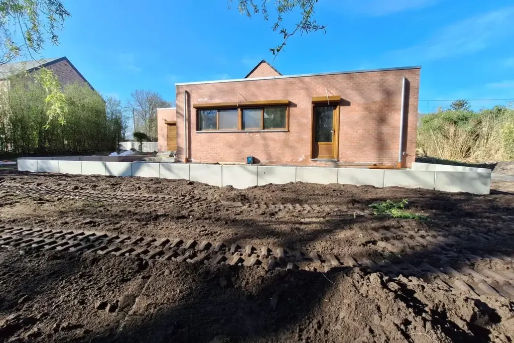 Rangée d'éléments de béton gris alignés au bord d'une plateforme devant une maison en briques, sur une terre retournée marquée de traces de chenilles