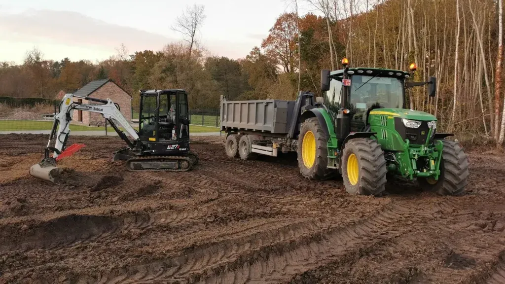 Mini-pelle et tracteur vert avec remorque benne sur un terrain de terre décapée, marqué par les traces de chenilles et de pneus