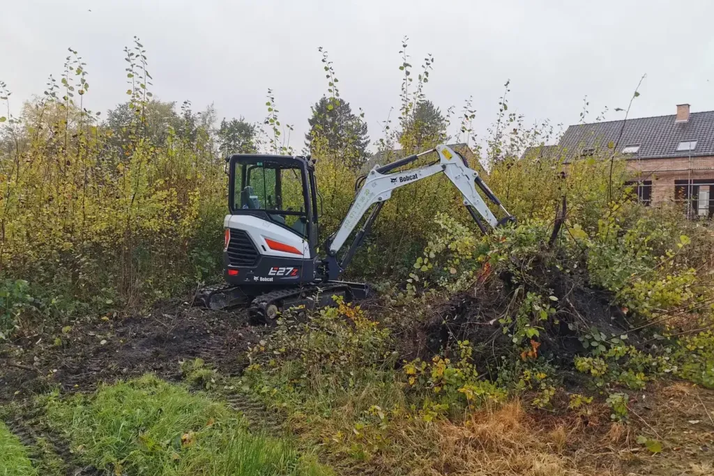 Mini-pelle blanche qui dégage un tas de ronces et de broussailles au bord d'un terrain en friche, avec une maison au fond à droite